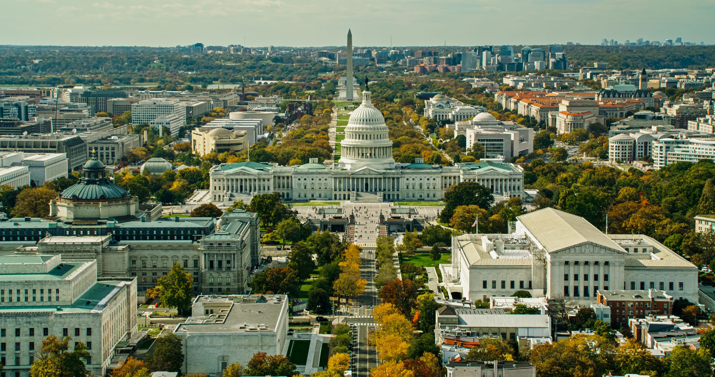 Helicopter shot of the Library of Congress, the Capitol, and the Supreme Court Building in Washington, D.C. on a hazy afternoon in Fall, with the Washington Monument and Lincoln Memorial beyond.